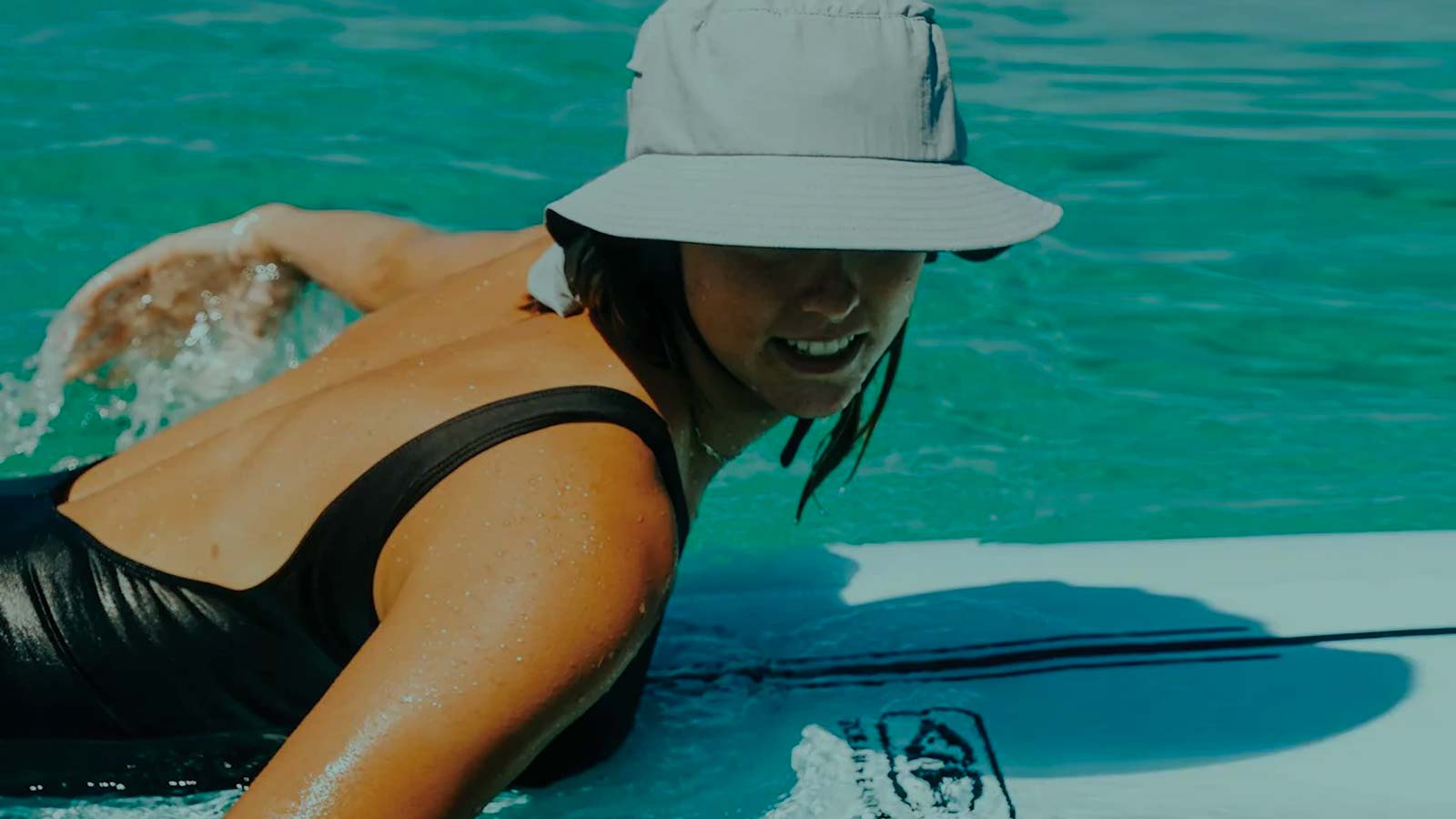 Girl paddling on a surfboard with a smile under the surf hat shade.
