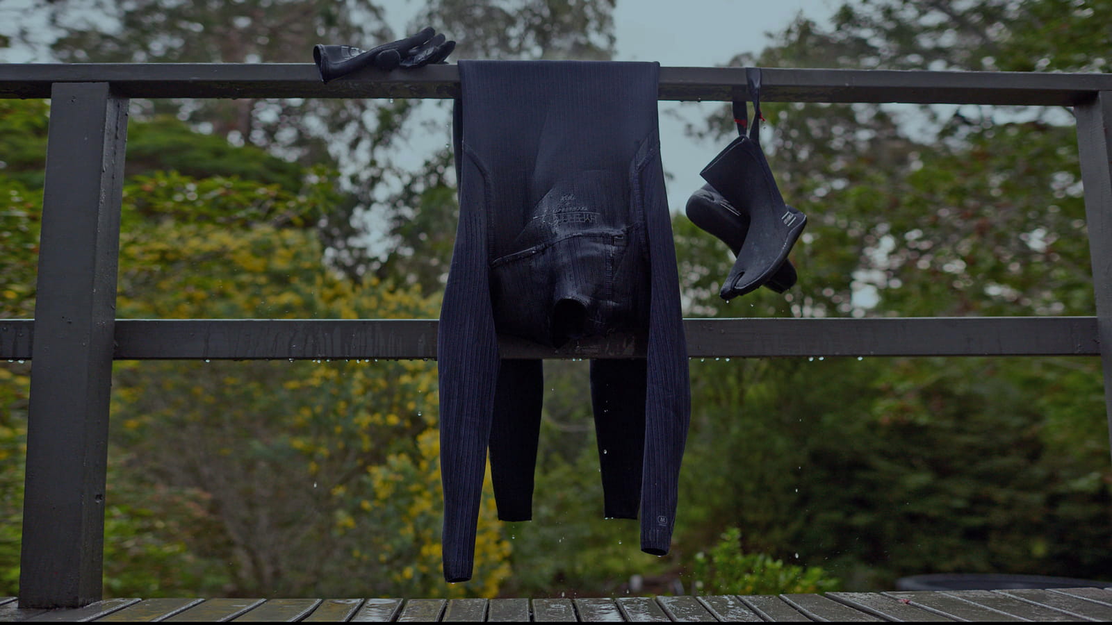 Wet wetsuit, neoprene gloves and neoprene boots are drying on the wooden railings. Black Neoprene Wetsuit. Green trees on the background.
